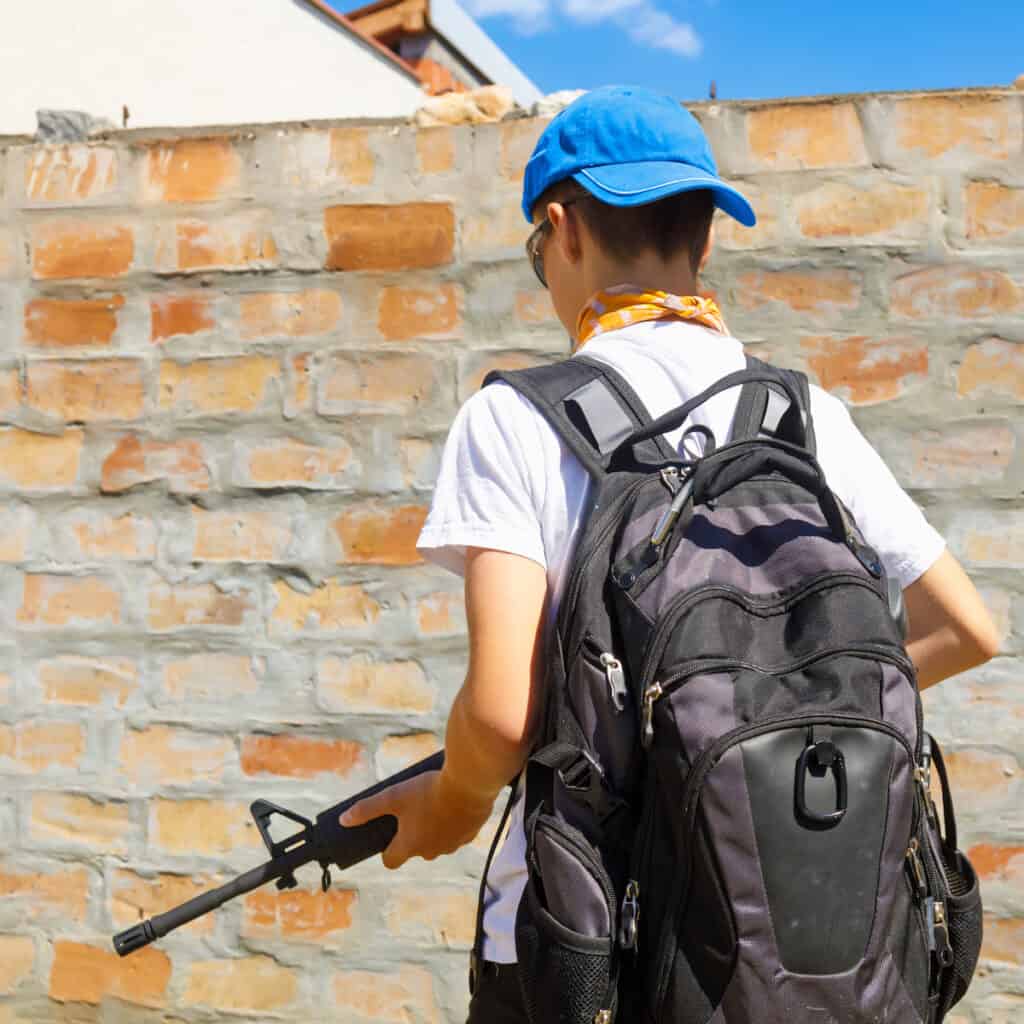 Photo of a young man holding an assault rifle, facing away, wearing a backpack