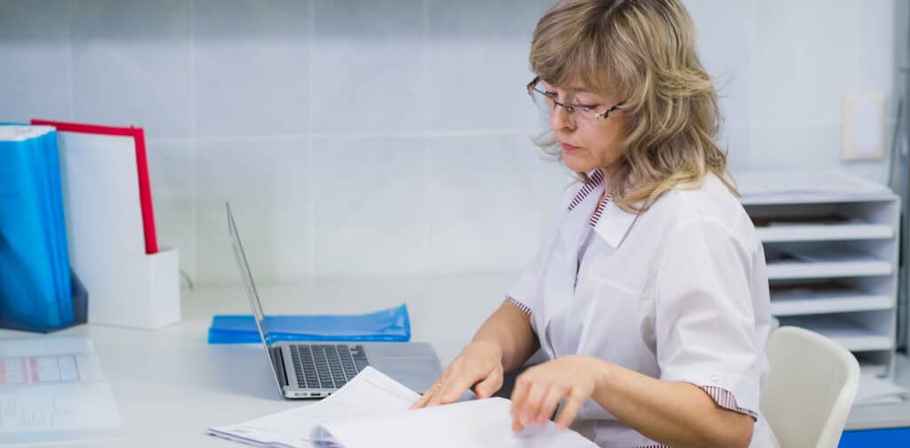 A woman sits at a desk in a hospital thumbing through papers near a laptop.