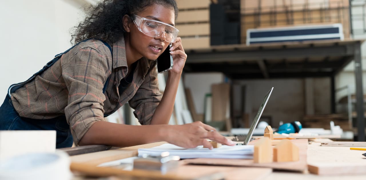 Young female carpenter using smart phone and working with laptop computer in wood workshop. Black female carpenter contact to customer by smart phone.